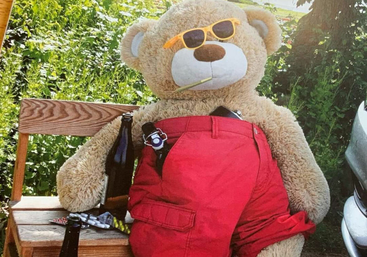 Ein großer Teddybär sitzt auf einer Bank und hat einen Joint im Mund und neben sich Bierflaschen stehen